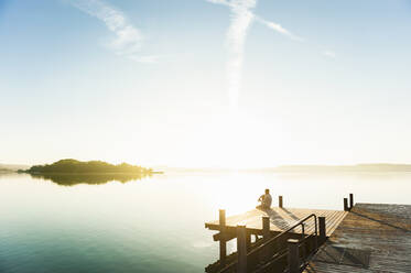 Young man sitting on jetty at lake enjoying sunrise - DIGF11949