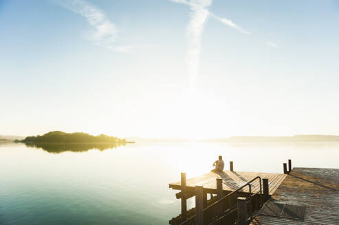 Junger Mann sitzt auf einem Steg am See und genießt den Sonnenaufgang - DIGF11949