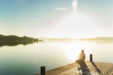 Young man sitting on jetty at lake enjoying sunrise - DIGF11950