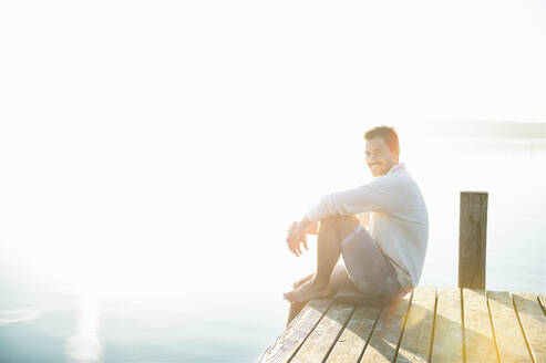 Porträt eines lächelnden jungen Mannes, der bei Sonnenaufgang auf einem Steg am See sitzt - DIGF11952