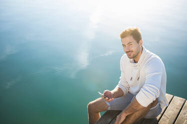 Portrait of smiling young man with smartphone sitting on jetty at lake - DIGF11953