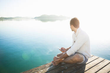 Junger Mann mit Smartphone sitzt auf Steg am See bei Sonnenaufgang und schaut in die Ferne - DIGF11954