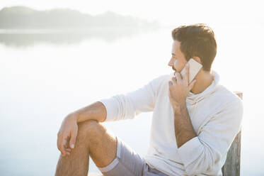 Lächelnder junger Mann am Telefon sitzt am See bei Sonnenaufgang und schaut in die Ferne - DIGF11955