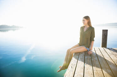 Young woman sitting on jetty at lake by sunrise - DIGF11956