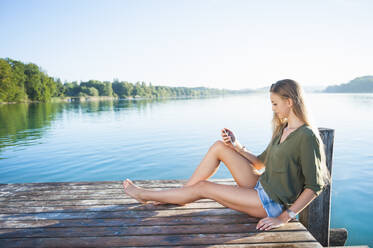 Junge Frau sitzt auf einem Steg am See und schaut auf ihr Handy - DIGF11958