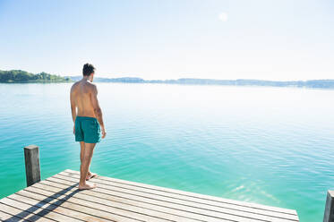 Back view of young man in swimming shorts standing on jetty looking at lake - DIGF11966