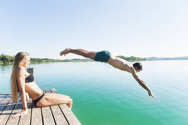 Junger Mann in Badeshorts springt vom Steg in den See, während seine Freundin ihn beobachtet - DIGF11970