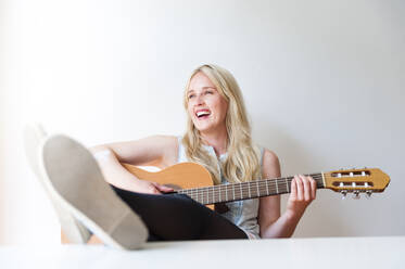 Portrait of laughing blond woman with feet up playing guitar - DIGF12010