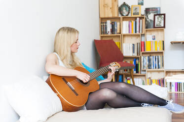 Blonde junge Frau sitzt auf der Couch zu Hause und spielt Gitarre - DIGF12014