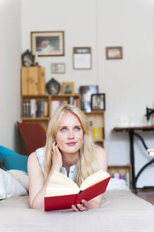 Porträt einer lächelnden blonden Frau, die mit einem Buch auf der Couch liegt und in die Ferne schaut - DIGF12015