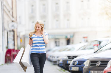 Portrait of happy blond woman with shopping bags looking at smartphone while walking on pavement - DIGF12025