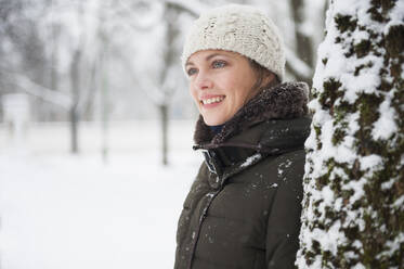 Portrait of smiling woman leaning at tree trunk in winter - DIGF12033