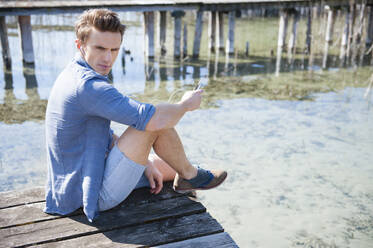 Gut aussehender Mann im mittleren Erwachsenenalter, der sein Smartphone benutzt, während er auf einem Pier über einem See sitzt - DIGF12037