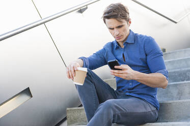 Businessman holding coffee using smart phone while sitting on steps - DIGF12050