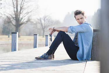 Thoughtful handsome mid adult man looking away while sitting on pier - DIGF12055