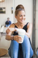 Portrait of laughing woman with bun sitting in a coffee shop looking out of window - DIGF12058