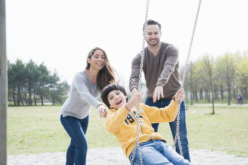 Glückliche Eltern, die ihren Sohn auf einer Schaukel im Park gegen den klaren Himmel schieben - DIGF12099