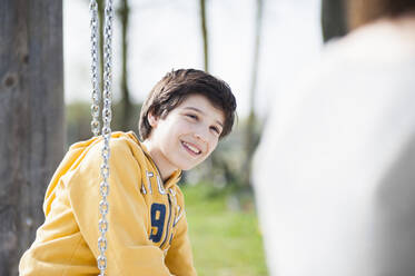 Happy boy looking at mother while sitting on swing in park - DIGF12101