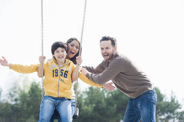 Cheerful man pushing mother and son on swing at park against clear sky - DIGF12102