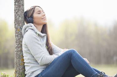 Relaxed woman listening music while leaning on tree trunk at park - DIGF12109