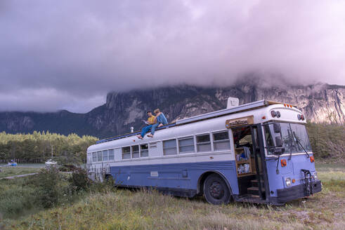 Zwei Menschen sitzen auf dem Dach eines Schulbusses und lesen ein Buch vor einem Berg - CAVF82265