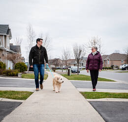 Ein Mann und eine ältere Dame gehen mit ihrem Hund auf dem Bürgersteig eines Vorstadtviertels spazieren. - CAVF82336