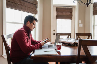 Mann mit Brille arbeitet von zu Hause aus an einem Computer am Esstisch. - CAVF82340