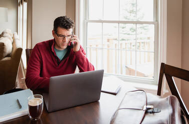 Man on cellphone working from home using a computer at a dining table. - CAVF82342