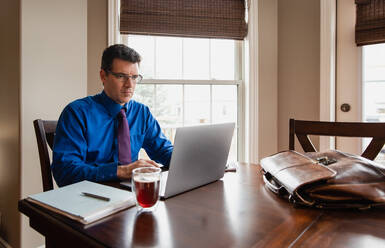 Mann in Hemd und Krawatte arbeitet von zu Hause aus am Computer am Esstisch. - CAVF82349