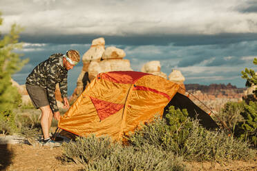 Camper in Camo-Windjacke baut in der Wüste von Utah sein Zelt auf - CAVF82466