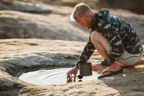 Mann in Tarnjacke holt Wasser zum Kochen aus einer flachen Pfütze - CAVF82505