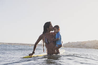Porträt einer Surfer-Mutter, die ihren Sohn neben einem Surfbrett im Meer hält - CAVF82544