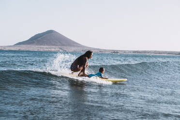 Gezogene Rückansicht von Mutter und Sohn beim Surfen auf einer kleinen Welle im Meer - CAVF82556