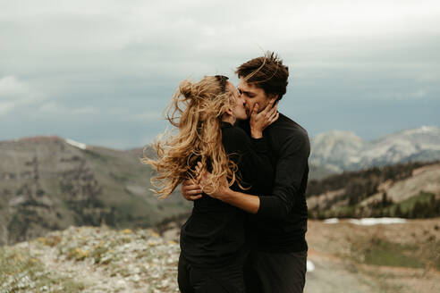 Neu verlobtes weißes Paar ganz in Schwarz küsst sich auf einem windigen Bergrücken - CAVF82716