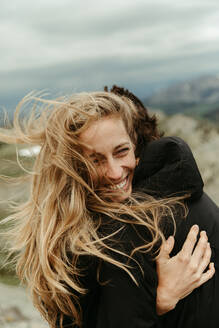 Frau mit windgefegten langen blonden Haaren lacht und umarmt ihren Partner - CAVF82721