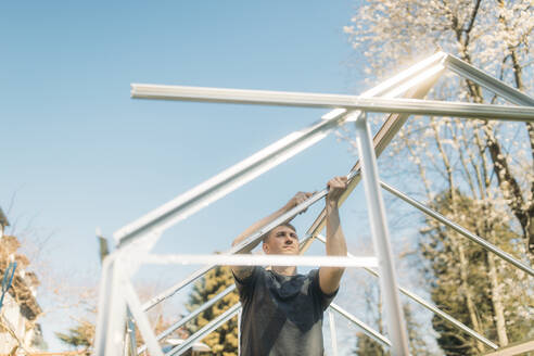 Mann beim Bau eines Gewächshauses im Garten an einem sonnigen Tag gegen einen klaren blauen Himmel - GUSF03777
