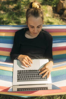 Junge Frau, die einen Laptop benutzt, während sie in einer Hängematte im Garten sitzt, während eines sonnigen Tages - GUSF03800