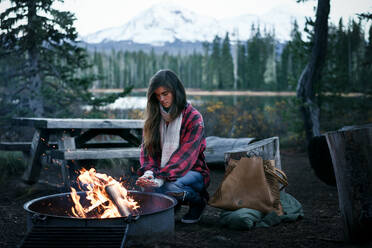 Young Woman crouching by fire warming hands - CAVF82830