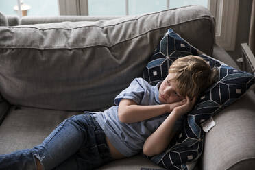 Trauriger blonder Junge sieht fern, während er auf der Couch liegt - CAVF82882