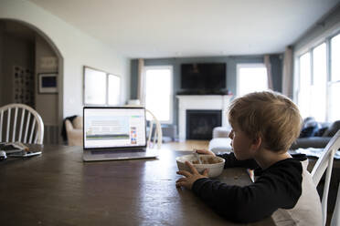 Wide Side View of Blonde Boy Eating Cereal Watching Lesson on Laptop - CAVF82888