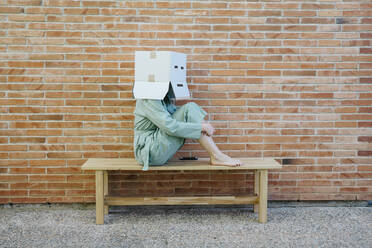 Woman sitting on bench in front of brick wall with serious face on cardboard box, hugging knees - AFVF06322