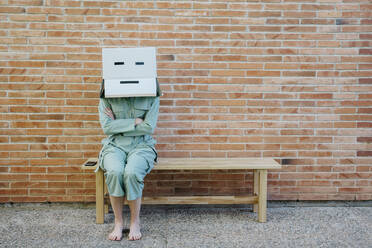Woman sitting on bench in front of brick wall with serious face on cardboard box, arms crossed - AFVF06323
