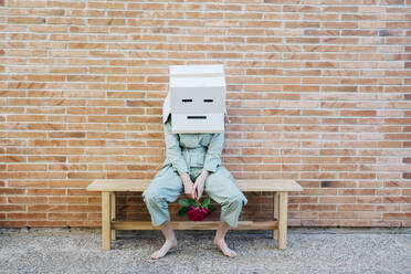 Disappointed woman sitting on bench in front of brick wall with sad face on cardboard box - AFVF06326