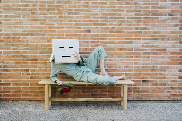 Bored woman with red rose, wearing cardboard box with sad face, lying on bench in front of brick wall - AFVF06327