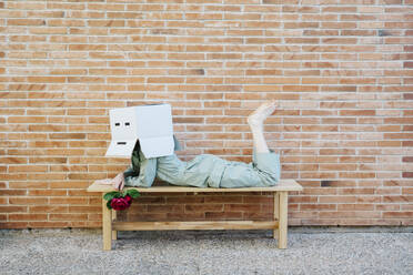 Bored woman with red rose, wearing cardboard box with sad face, lying on bench in front of brick wall - AFVF06328