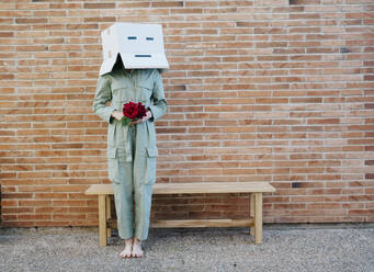 Woman holding red rose, wearing cardboard box with happy face, standing by bench in front of brick wall - AFVF06331