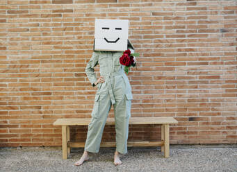 Woman holding red rose, wearing cardboard box with happy face, standing by bench in front of brick wall - AFVF06333