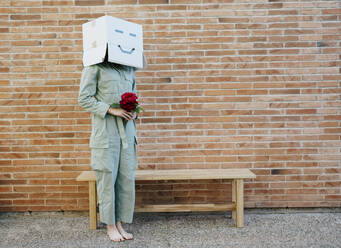 Woman holding red rose, wearing cardboard box with happy face, standing by bench in front of brick wall - AFVF06334