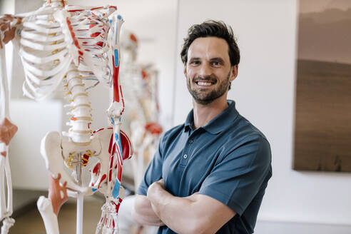 Physiotherapist standing by anatomical skeleton with arms crossed - DAWF01547