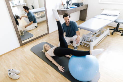 Physiotherapeutin hilft einer Patientin, die mit einem Fitnessball übt, auf einer Gymnastikmatte liegend - DAWF01577